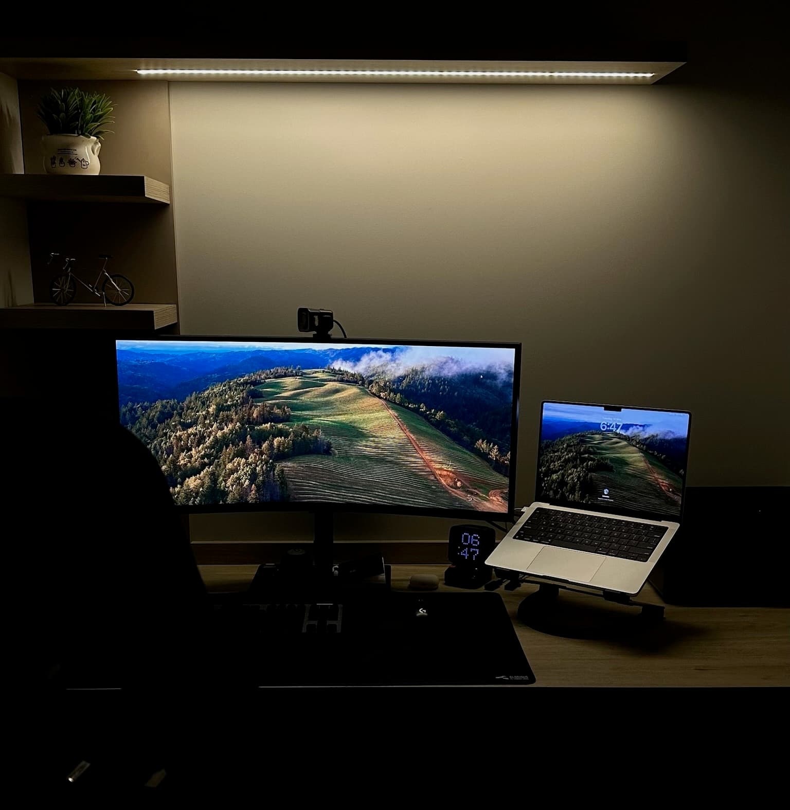 Escritorio de noche bajo la barra LED cálida. Ultrawide curvo con wallpaper de montañas, MacBook en stand, reloj Ditoo chico, teclado mecánico, luz cálida.